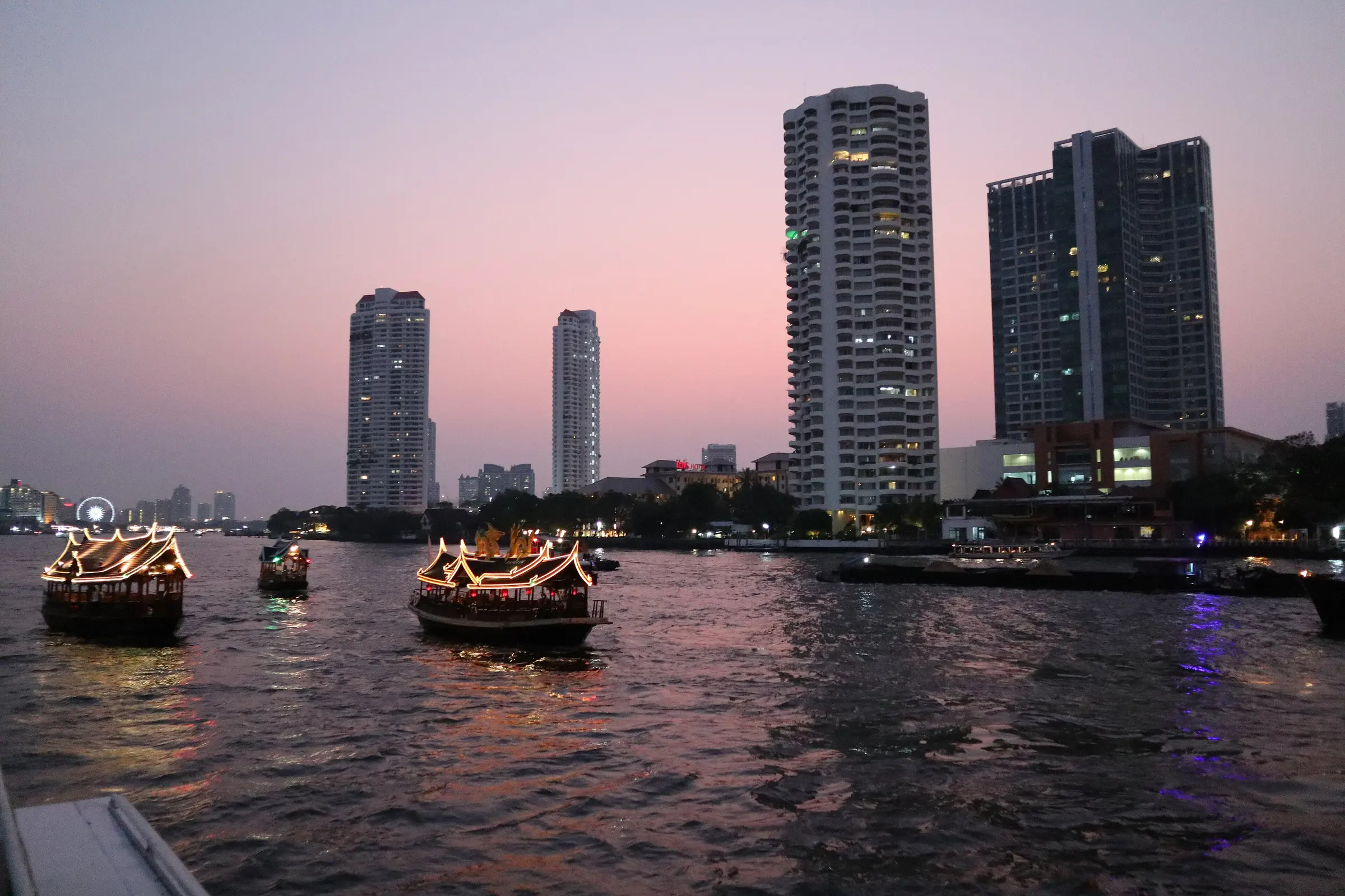 Boten op de Chao Phraya in Bangkok bij zonsondergang, met flats aan de oever