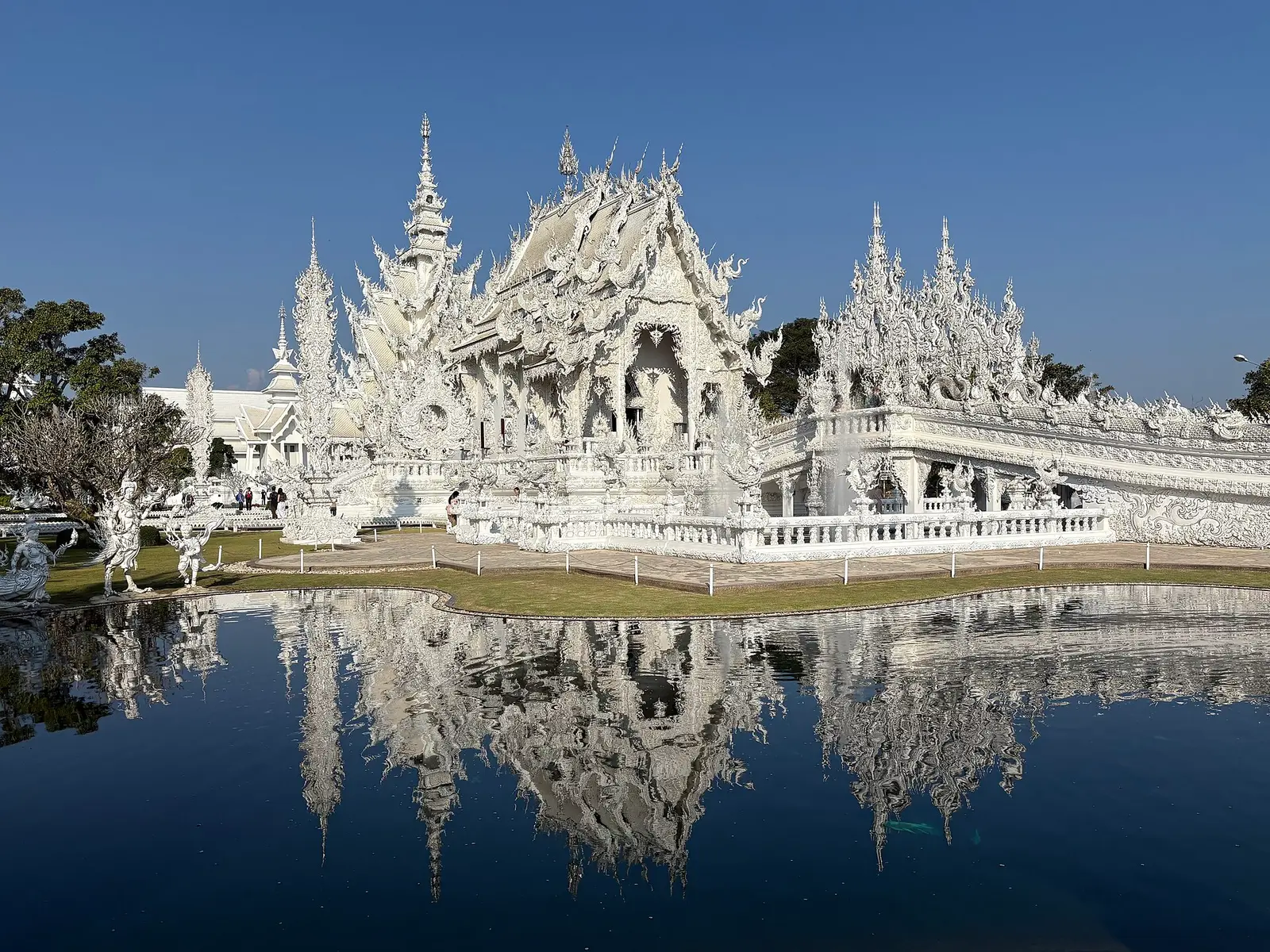 De witte tempel Wat Rong Khun in Chiang Rai, weerspiegeld in een donker watervlak onder een blauwe hemel