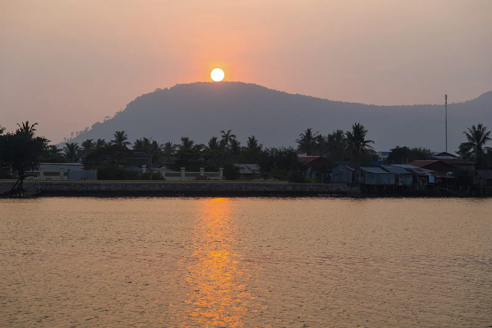 Zonsondergang boven een berg aan de rivier Praek Tuek Chhu bij Kampot, met huizen en palmen aan de oever