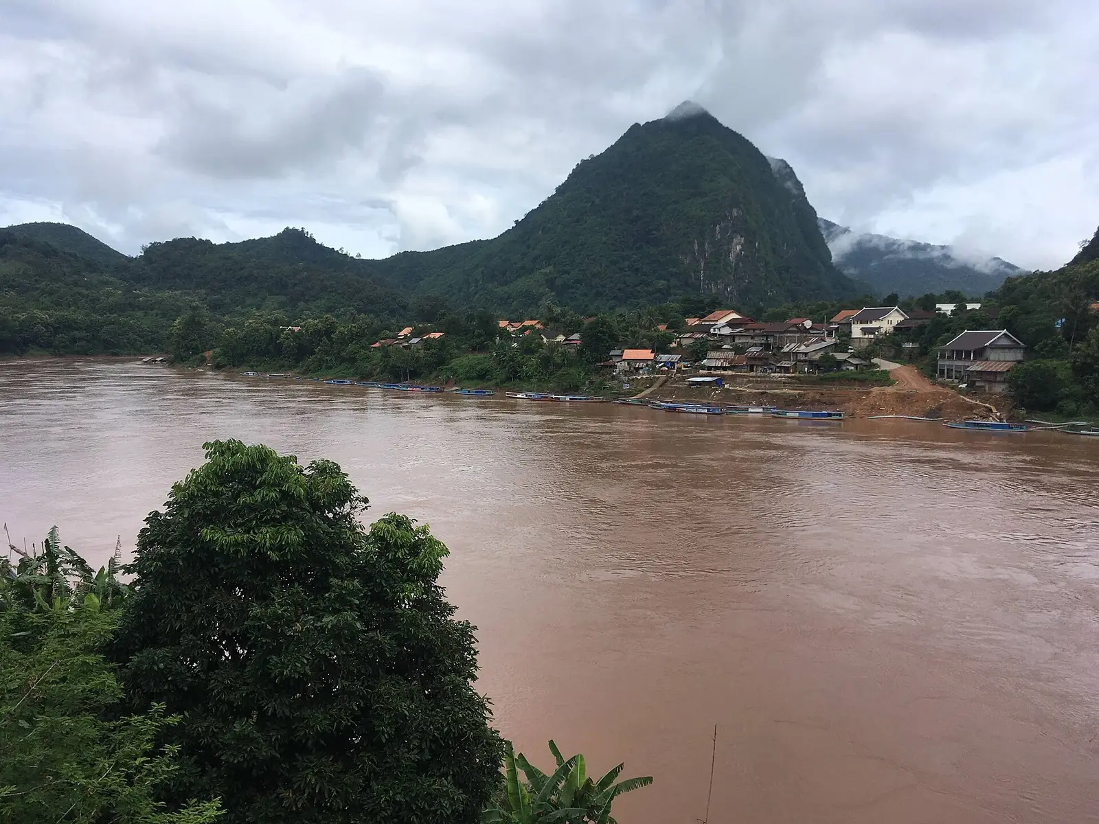 Houten huizen en boten aan de Nam Ou in Nong Khiaw, met kalksteenwanden erachter