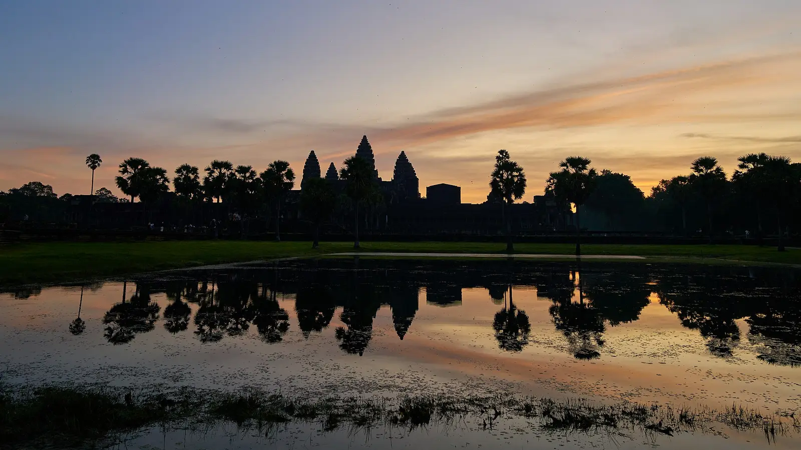 Angkor Wat tegen een oranje ochtendhemel, met de torens en palmen weerspiegeld in het water ervoor