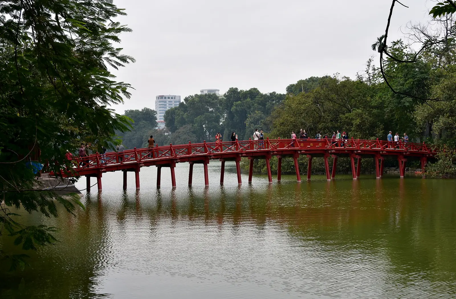 De rode Huc-brug over het Hoan Kiem-meer in Hanoi, met wandelaars en bomen eromheen